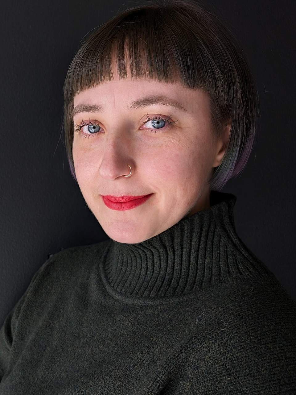 A person with blue eyes, short brown/blue/purple hair with bangs smiles at the camera wearing red lipstick. They have on a forest green mock neck sweater. The background is dark grey.